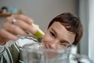 Portrait of boy smiling while squeezing lemon slice into glass pitcher, focusing on hand movement...