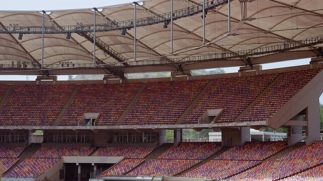 empty stadium grandstand under sweeping canopy wide panoramic view of tiered red seats and steel truss roof, maintenance crew inspecting concourse, event manager surveying sightlines, late afternoon