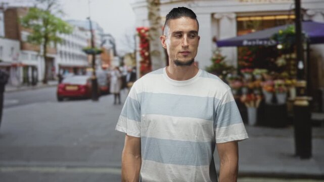 Man gazes sideways at flower stall, striped t shirt and bare forearm visible, standing on a busy street; quiet contemplation.