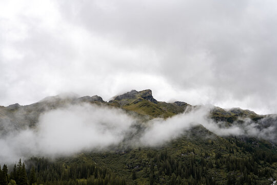 Low clouds and mist drift across a rugged mountain ridge above dense evergreen forest, creating a moody and atmospheric alpine landscape.