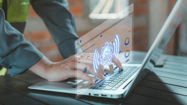 Industrial engineer typing on laptop with virtual smart gear and sensor signal hologram for equipment monitoring, predictive maintenance, OEE Overall Equipment Effectiveness, Efficiency Safety