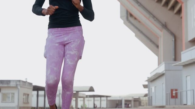 woman running toward camera with closeup leg action in purple leggings and sneakers, athletic form and breathing visible during energetic sprint near warehouse, confident athlete testing speed