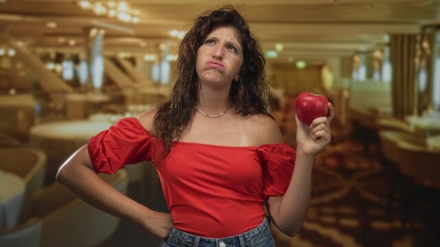 Young hispanic woman holding red apple and grimacing in restaurant dining room, wearing off shoulder blouse; disgust reluctance.