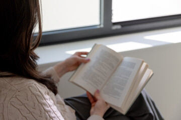 Naklejka premium Real woman sitting comfortably by a bright window, absorbed in reading a book, enjoying slow living, everyday comfort and the simple pleasure of appreciating quiet moments.