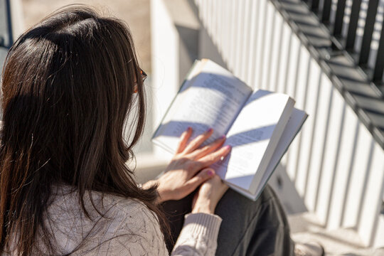 Real woman sitting on outdoor steps in warm sunlight, relaxed and absorbed in reading a book, enjoying slow everyday life, real moments and simple quiet comfort.