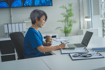 Senior Female Doctor Writing Notes While Holding Coffee in Clinic Office