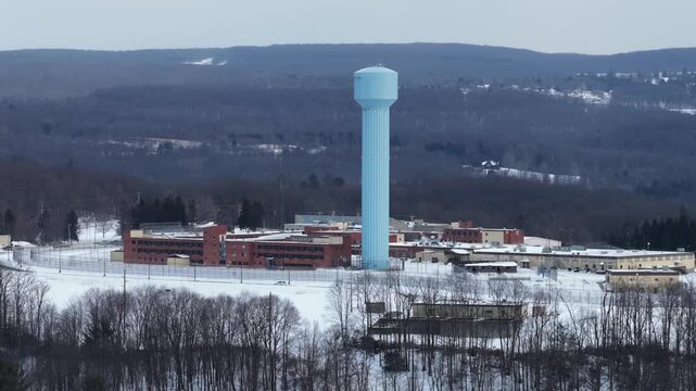 Aerial view of a correctional facility in Loretto, Pennsylvania, surrounded by snow-covered grounds and forested hills, with tall blue water tower rising above complex. Approaching shot. Pa, USA.