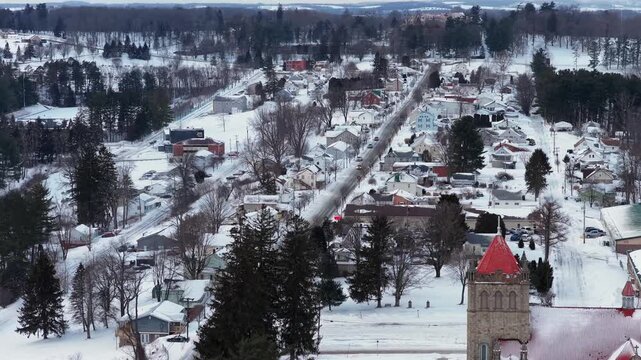 Aerial winter view of Loretto, Pennsylvania, with snow-covered Main Street, small homes and church with red roof surrounded by trees in quiet rural town. Wide shot. Peaceful scene in USA.