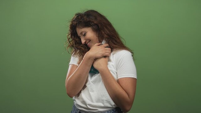 Woman hugging a teal cold pack to chest with clasped hands and a gentle smile against a green studio wall; contentment.