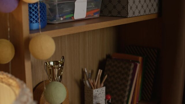 Close up of wooden shelves with different boxes full of notebooks and stationery, books, models of molecules and shiny golden goblet in teenage room
