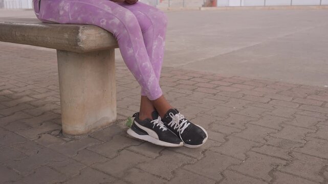 black woman legs seated on bench, purple patterned leggings and black white sneakers on textured pavement, closeup of feet and lower legs, concrete urban plaza setting, relaxed waiting pose