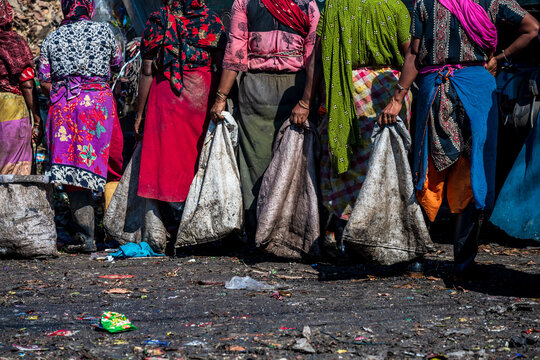 Chattogram, Bangladesh - 26 November 2025: View of women in vibrant saris holding sacks, standing on dark, uneven ground strewn with scattered debris and remnants of discarded materials.
