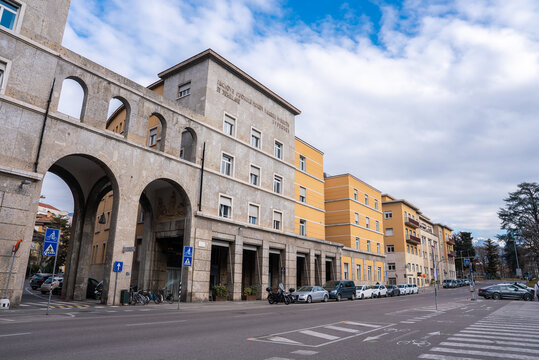 Street level view in Bolzano, Italy shows rationalist arcades, arches, stone facade with inscription and relief, mustard yellow blocks, parked cars, bikes, zebra crossings.