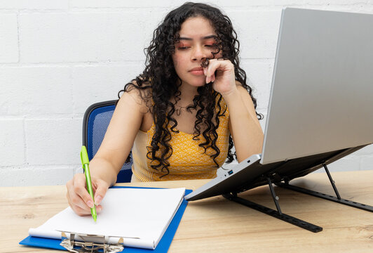 Young woman sitting at her desk next to her computer, writing notes on work papers. Concept of productivity, workplace organization, and professional focus