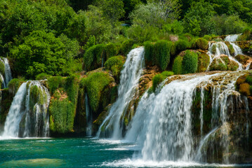 Obraz premium Stunning multi-tiered waterfall cascading from green, moss-covered rocks, surrounded by lush vegetation and dense forest, creates serene natural landscape. Krka National Park is in Croatia.