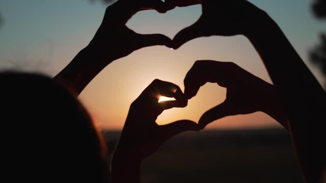 Hands form heart shape at sunset beach. Couple makes love gesture with hands toward sun. Romantic silhouette shows heart symbol. Love hands create heart at sunset. Beach romance with heart gesture.