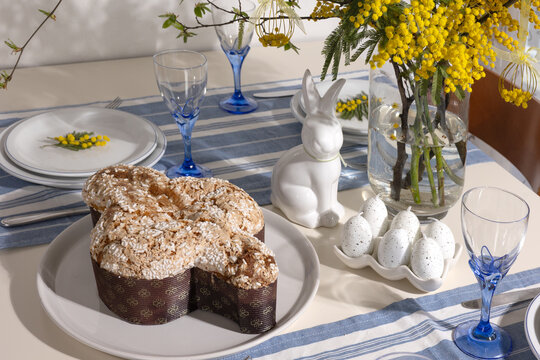 Easter table decorated with colomba di pasqua cake, decorative bunny, speckled egg candles, blue glassware, and vibrant mimosa branches in a vase, preparing for a festive spring celebration