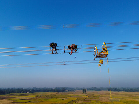 Aerial view of technicians suspended high on power lines against a clear blue sky, a testament to human endeavor, Joypurhat, Rajshahi Division, Bangladesh.