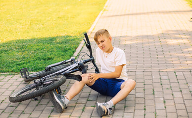 Teenager kid boy fell off a bicycle and injured his knee outdoors in park. Child sitting down on the road.