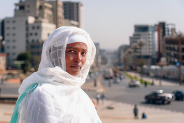 Close-up portrait of an Ethiopian woman wearing a traditional white Habesha kemis with a delicate green-bordered netela, set against the vibrant, sun-drenched backdrop of a modern Addis Ababa street.