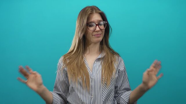 Young blonde woman raises both hands with palms facing forward in studio against teal backdrop; indifference.