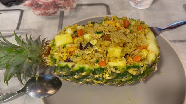 Hand uncovers lid of traditional Thai fried rice served inside a hollowed pineapple, mixed with egg, vegetables, and spicesin a casual dining setting