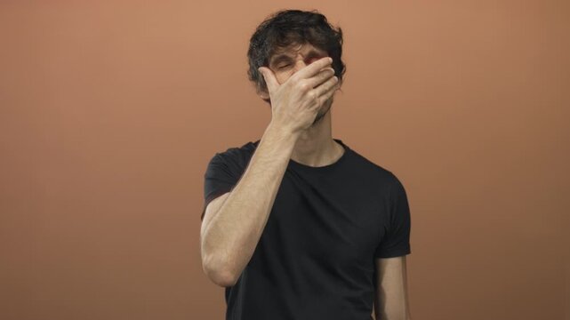 Young man places hand on forehead and shades eyes with palm against a tan studio wall; concern introspection.