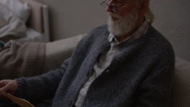 Tilt up slowmo shot of concerned elderly Caucasian man in eyeglasses and knitwear sitting alone in dimly lit room and looking at overdue bills, struggling with financial hardship