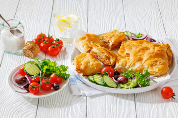 Sliced savory greek meat bougatsa of crispy phyllo pastry with minced meat filling on a plate with cucumbers, tomatoes, kalamata olives, red onion, parsley on white wooden table, not AI generated