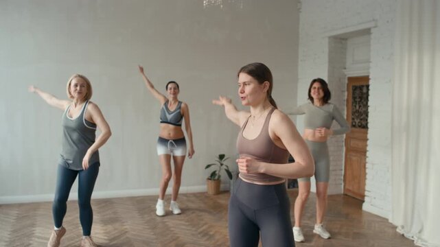 Group of women doing dancing cardio warm up with instructor before yoga class in spacious bright fitness studio room