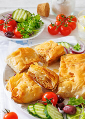 Bougatsa phyllo pastry with minced meat on white plate with red onion, olives and cherry tomatoes, cucumbers, Greek cuisine, vertical view, close-up, not AI generated