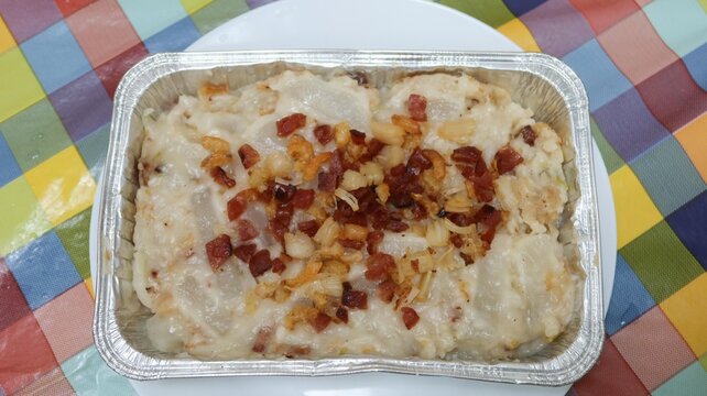 close up of Hong Kong traditional style homemade Chinese steamed turnip cake topped with Chinese sausage, dried shiitake mushrooms and dried shrimp in a Foil container on a white plate over colorful c
