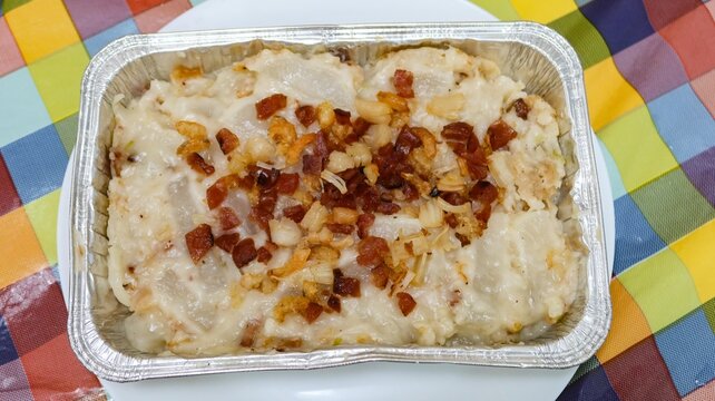 close up of Hong Kong traditional style homemade Chinese steamed turnip cake topped with Chinese sausage, dried shiitake mushrooms and dried shrimp in a Foil container on a white plate over colorful c