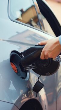 Close-up. Woman plugs in charger into socket of her modern new electric car. Girl in a casual summer outfit, charging a black electric car parked on the street. Slow motion.