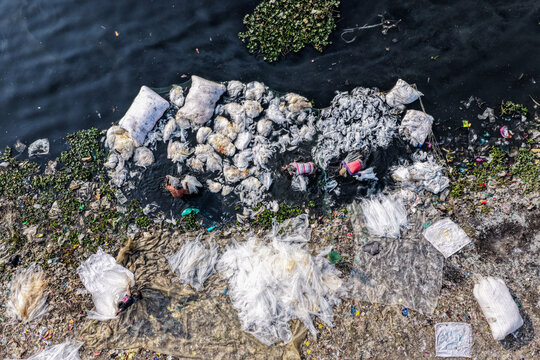 Aerial view of the riverbank choked with piles of discarded white fabrics and waste, contrasting with the dark water, Buriganga River, Dhaka, Dhaka Division, Bangladesh.