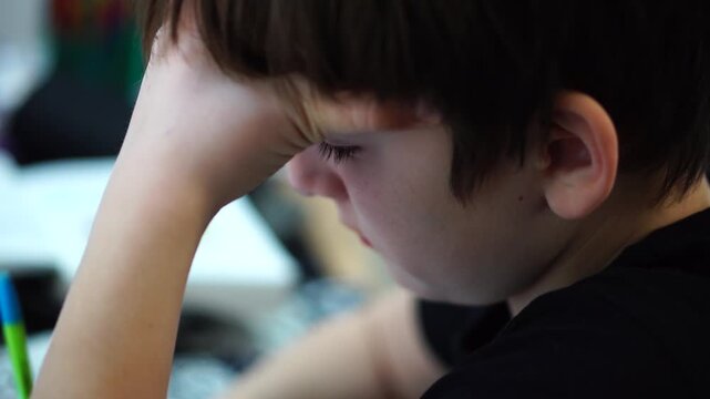Focused schoolboy resting head on his arm and writing notes, tired student doing homework.