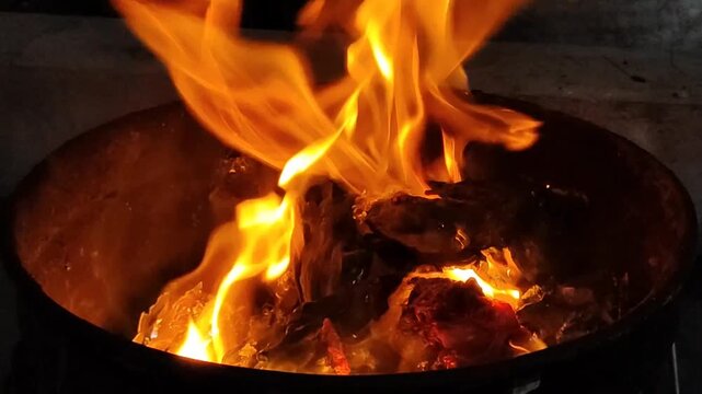 Close-up of glowing embers and bright flames rising from burning wood inside a dark metal fire pit, capturing intense heat, texture, and dramatic light in a rustic setting