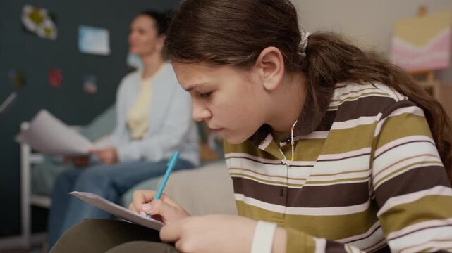 Close up of attentive teen girl sitting in bean bag chair and doing math test while her mother and tutor having conversation in background