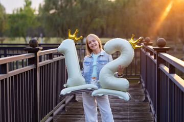 Smiling girl holding number 12 balloons on a bridge at sunset