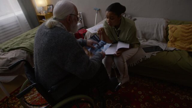 High angle handheld shot of senior Caucasian man in wheelchair sitting in dim lit bedroom holding brochure and asking questions to young Black female worker about social assistance programs