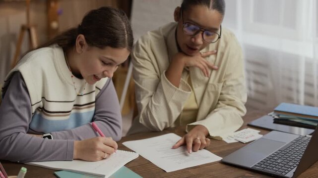 Medium shot of math tutor explaining study task, gesturing gently while teen student listening to her carefully and taking notes