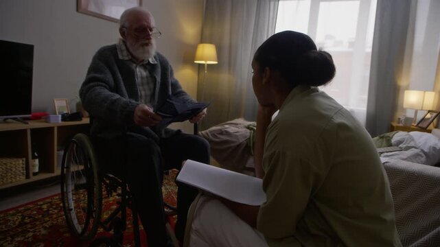 Side view shot of young African American female social worker helping elderly Caucasian man in wheelchair reading brochure during home care visit, explaining benefits of social service program