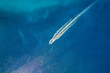 High-angle aerial shot of a single powerboat creating a sharp white wake through deep blue water, capturing the essence of speed, travel, and tranquility on a vast, sunlit open ocean surface. © _jure