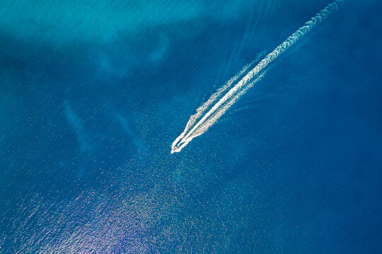 High-angle aerial shot of a single powerboat creating a sharp white wake through deep blue water, capturing the essence of speed, travel, and tranquility on a vast, sunlit open ocean surface.