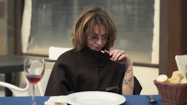 Stylish woman wear sunglasses and brown coat, vaping an electronic cigarette while use smartphone at restaurant table with glass of red wine.Concept of digital lifestyle, remote work