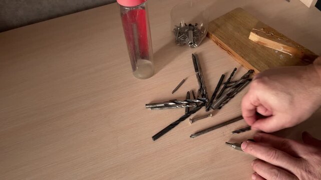 Hands sorting drill bits on wooden table selecting metal tools for drilling work workshop organization hardware equipment preparation