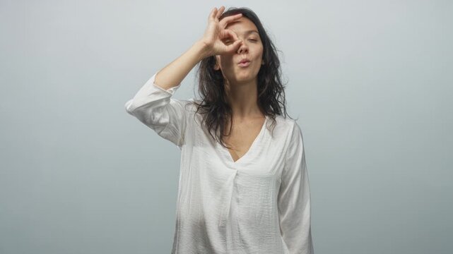 Woman in white blouse making ok sign with hand over eye, peering through finger circle and tilting head slightly in a neutral studio setting; playful curiosity.