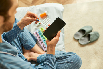 Handsome man in cozy home setting checks smartphone while managing medication routine thoughtfully