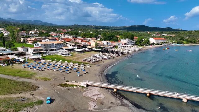 Aerial drone flyover of wooden pier and swimming area in Roda beach North Corfu Greece with tourists enjoying water sports and sunbathing on a sunny summer day