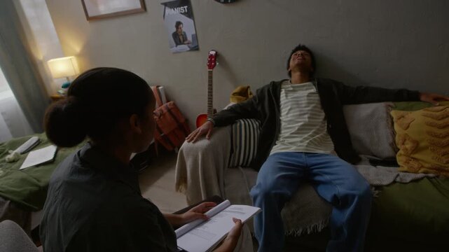 Medium long slowmo shot of gen Z Black boy sitting on couch in relaxed pose in messy room and answering questions of young female social worker, avoiding eye contact during home care visit